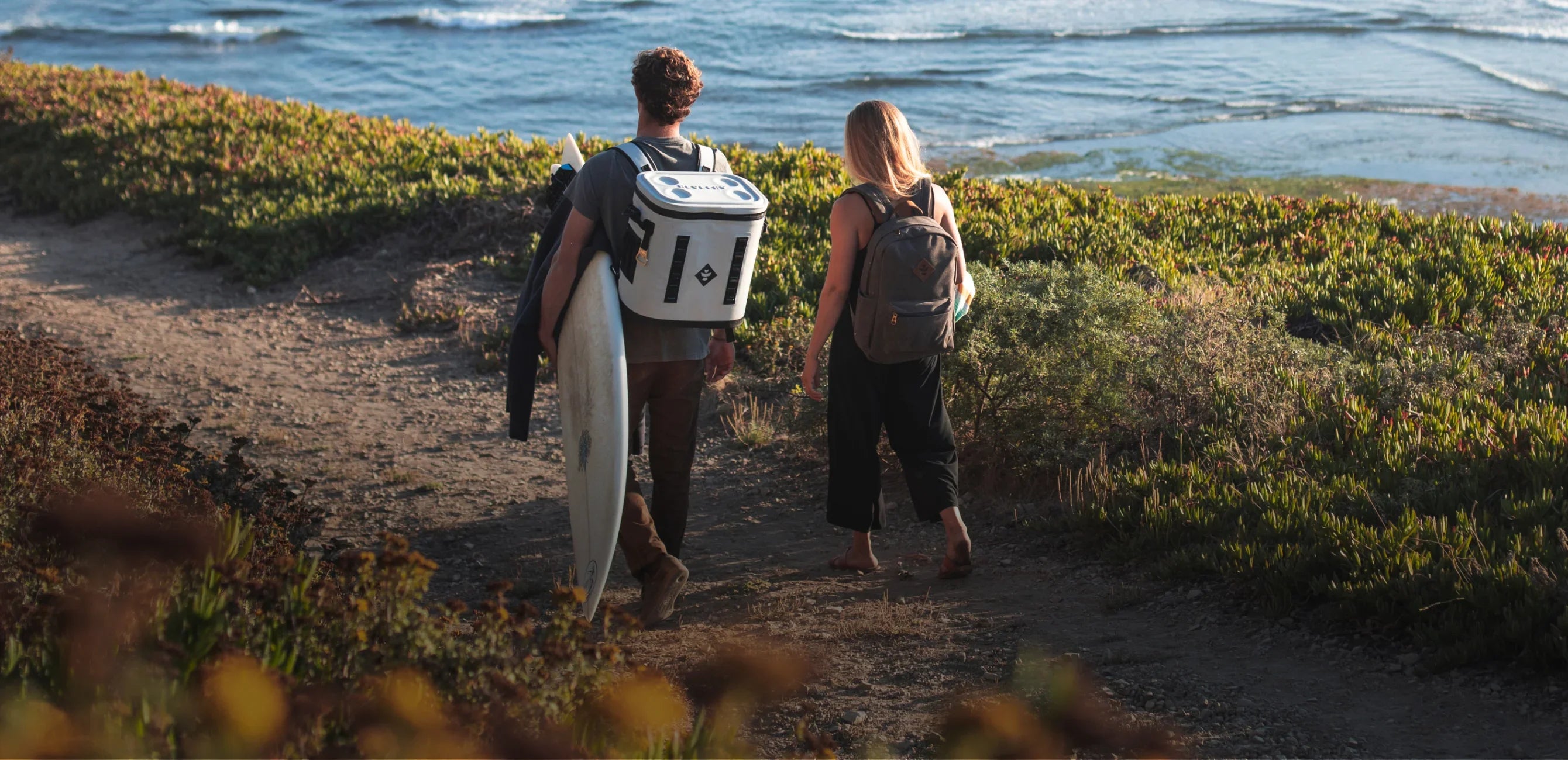 Two people walking along a path near the ocean with surfboards and backpacks.